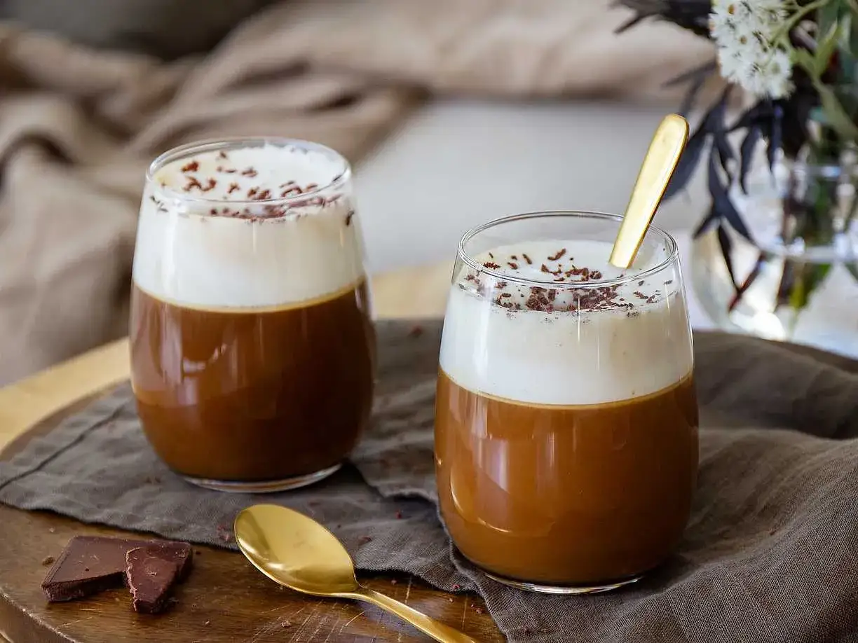 Två glas med krämig kaffe Karlsson, toppad med vispgrädde och chokladströssel. En gyllene sked vilar i det ena glaset.