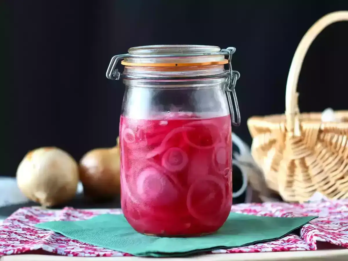 Frasco de vidrio con cebolla encurtida, rodajas rosadas vibrantes sumergidas en l&iacute;quido. Dos cebollas crudas al lado.