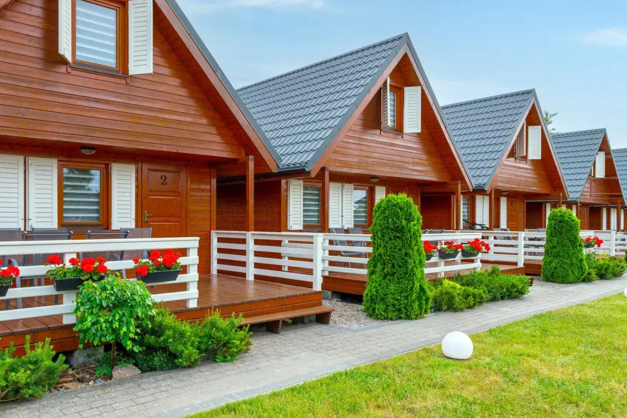 Urokliwe ustka domki nad morzem z drewnianymi fasadami i białymi balustradami, otoczone zielenią i kwitnącymi czerwonymi kwiatami.