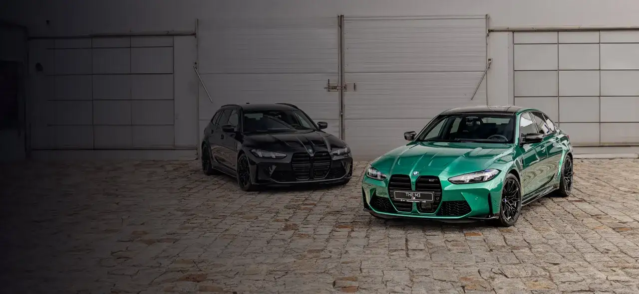 Dwa sportowe BMW X, jeden czarny, drugi zielony, stoją na brukowanym placu przed garażami.