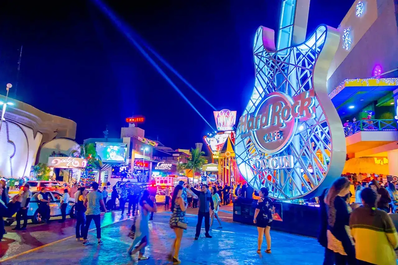 A vibrant night scene at Hard Rock Cafe Cancun, with a giant guitar sculpture and neon lights. People are enjoying the lively atmosphere, a perfect spot for gay Cancun nightlife.