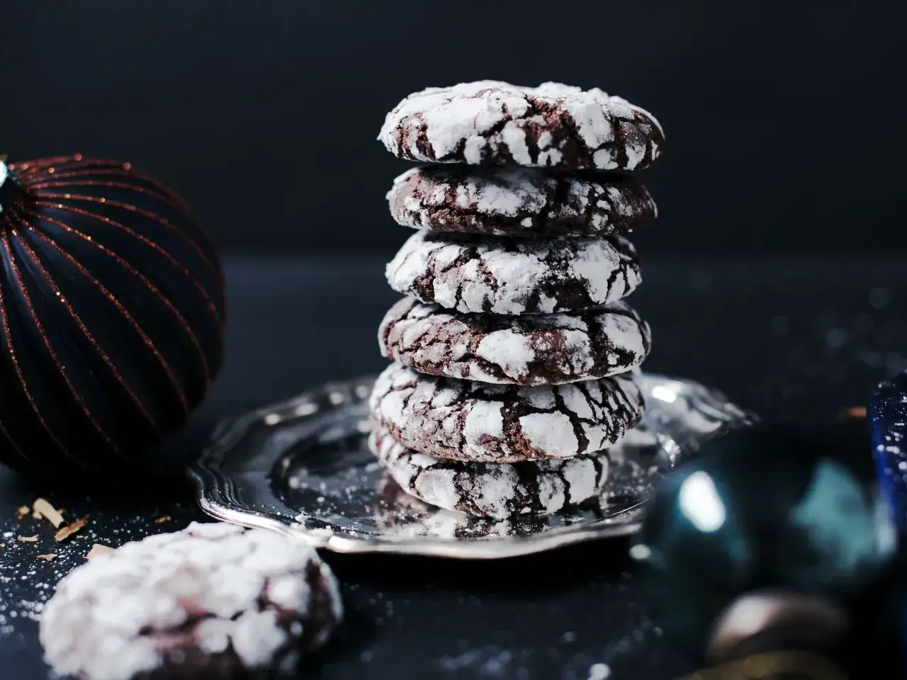 Gestapelte american cookies mit Puderzucker best&auml;ubt auf einem Silbertablett, daneben ein dunkler Weihnachtsbaumschmuck.