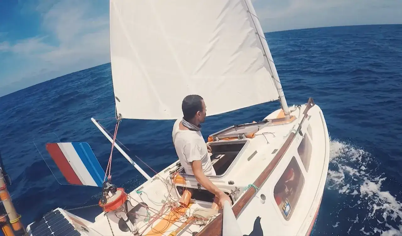 Un homme seul sur son petit bateau, le baluchon prêt pour l'aventure. Le vent gonfle la voile blanche sur l'océan bleu.