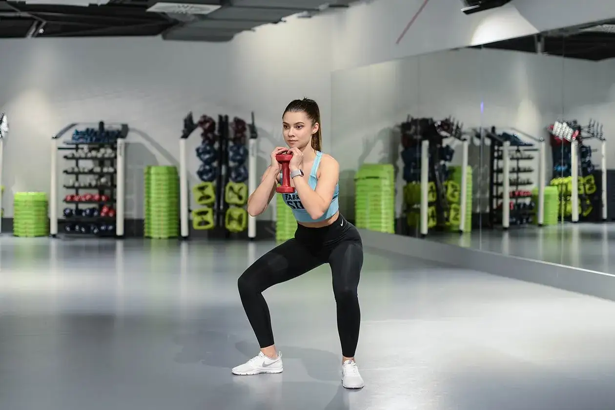 Kobieta wykonuje goblet squats z czerwoną hantlą w siłowni, w tle stojaki ze sprzętem.