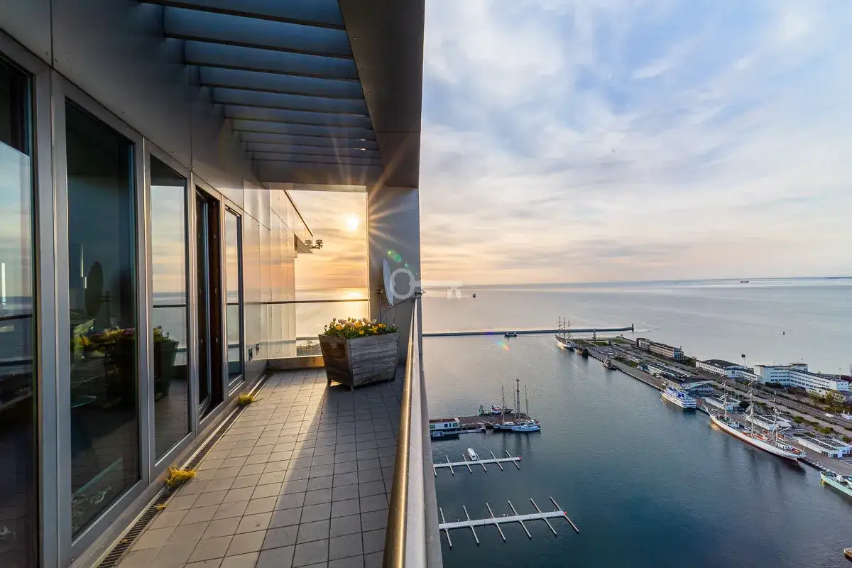 Widok z balkonu na port i morze o zachodzie słońca. W oddali widać żaglowce i inne statki, a także nowoczesne **sea towers**.