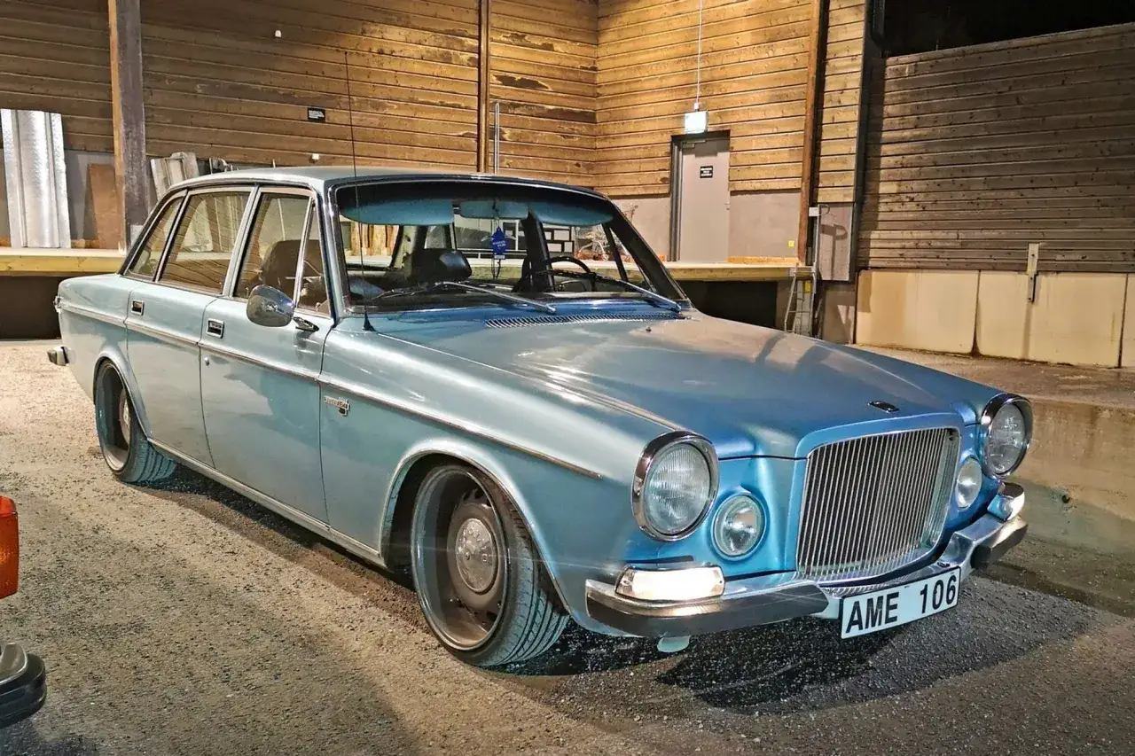 En ljusbl&aring; Volvo 164 fr&aring;n 1973 st&aring;r parkerad framf&ouml;r en tr&auml;fasad. Bilen har s&auml;nkts och har stilrena f&auml;lgar.
