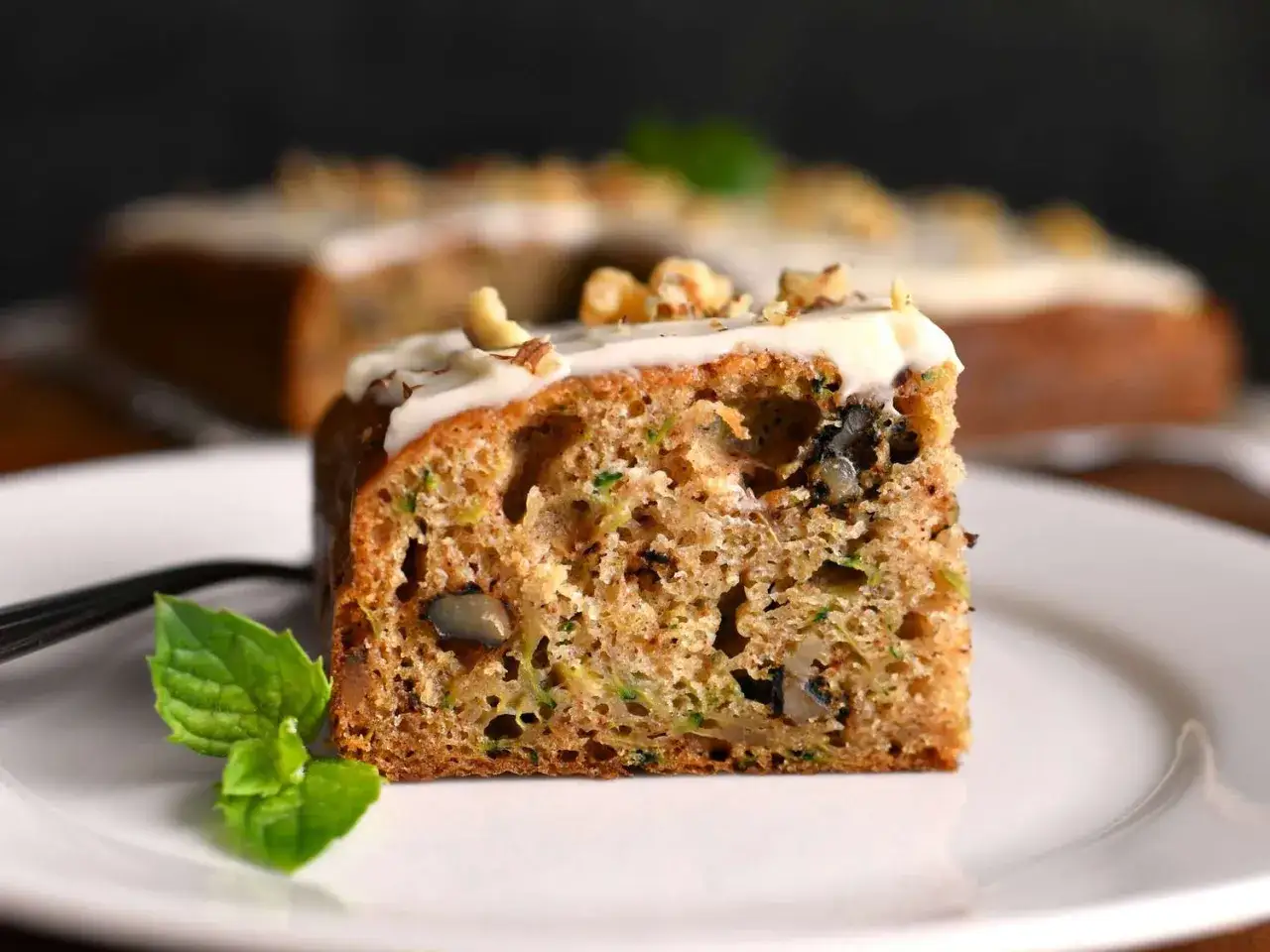 Ein saftiges St&uuml;ck Zucchini Kuchen mit N&uuml;ssen und einem cremigen Topping, garniert mit Minze.