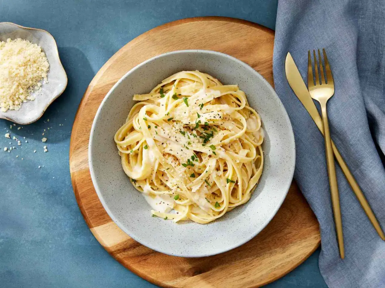 Miseczka fettuccine z kremowym, oryginalnym sosem alfredo, posypana parmezanem i natką pietruszki. Obok złote sztućce i ser.