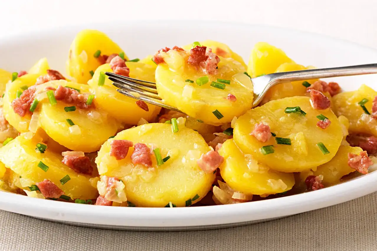 Ein Teller mit Kartoffelsalat mit Speck und Schnittlauch. Die Kartoffelscheiben sind goldgelb und mit kleinen Speckwürfeln und Zwiebeln garniert.
