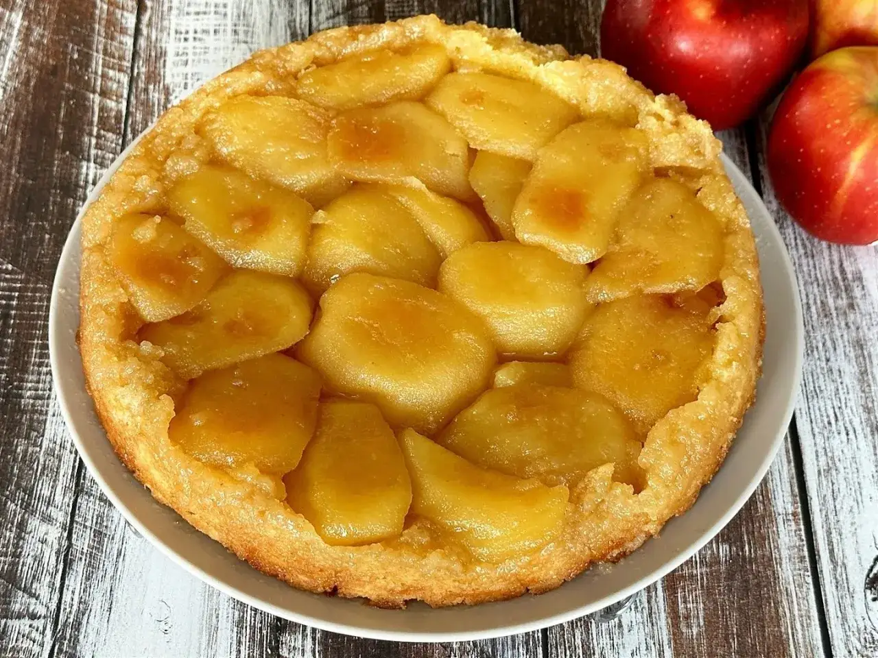 Pyszna tarta tatin z jabłkami, której owoce pod kruszonką kuszą złotym kolorem. Obok leżą czerwone jabłka.