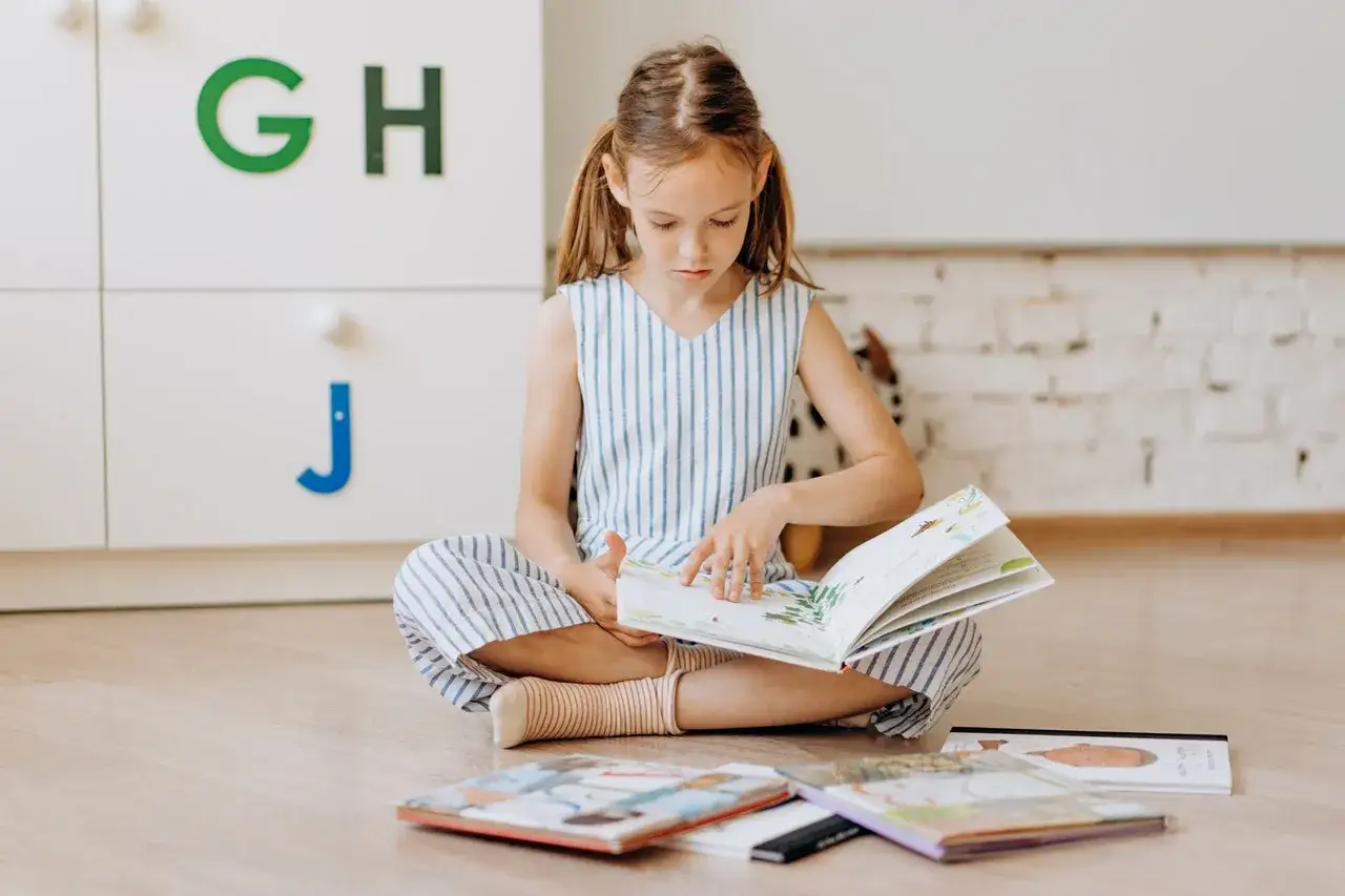 Ni&ntilde;a concentrada en la comprensi&oacute;n lectora 2 primaria, sentada en el suelo con libros. Las letras G, H y J decoran un mueble.
