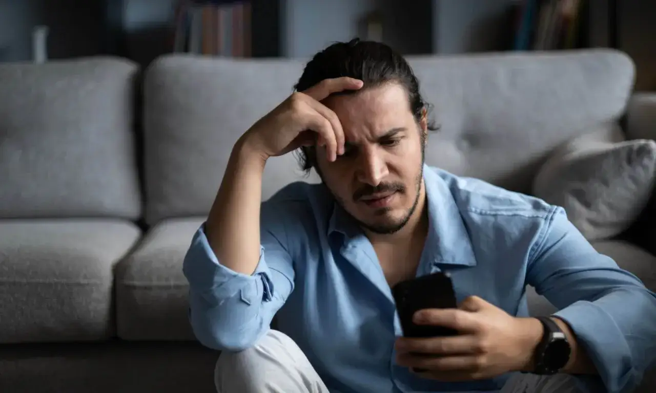 Uomo pensieroso con mano sulla fronte, guarda il telefono. La psicologia del chiudere il telefono in faccia è complessa, spesso legata a frustrazione o rabbia.