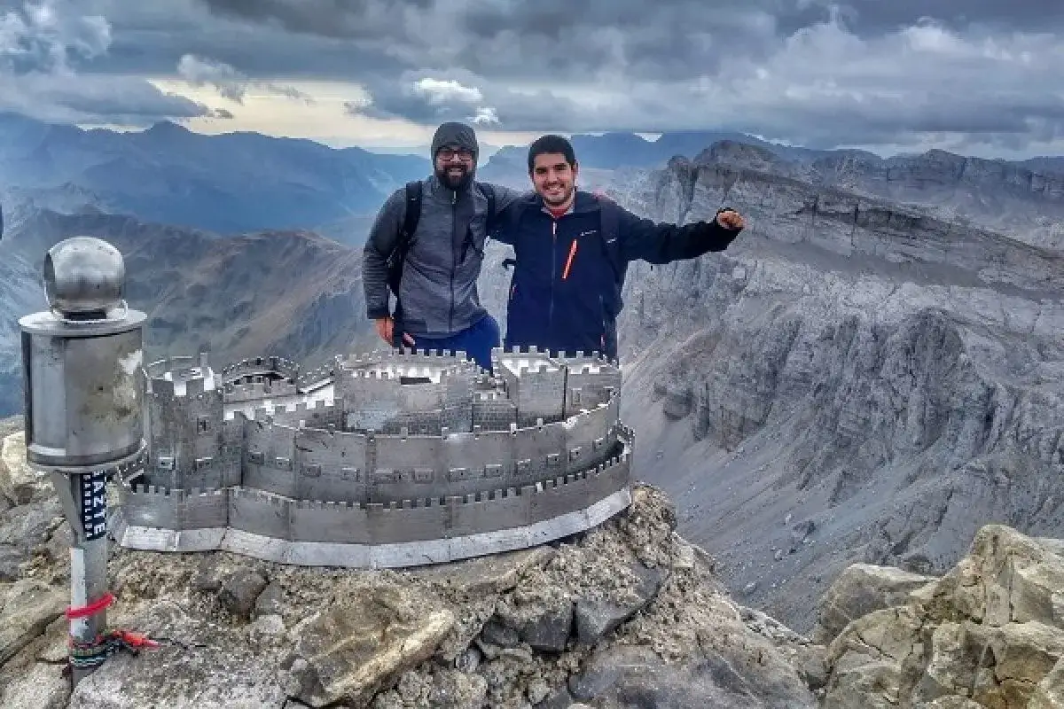 Dos amigos posan junto a una maqueta de un castillo en la cima de una montaña, con un paisaje montañoso espectacular de fondo. El **erregeen mahaia** parece un tesoro en este lugar remoto.