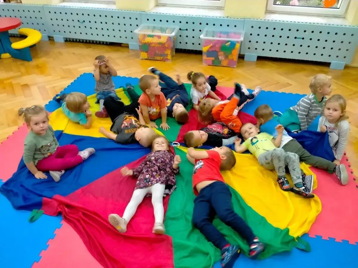 Dzieci leżą na kolorowym materiale, ciesząc się **zabawami na dzień dziecka**. Radość i beztroska maluchów.