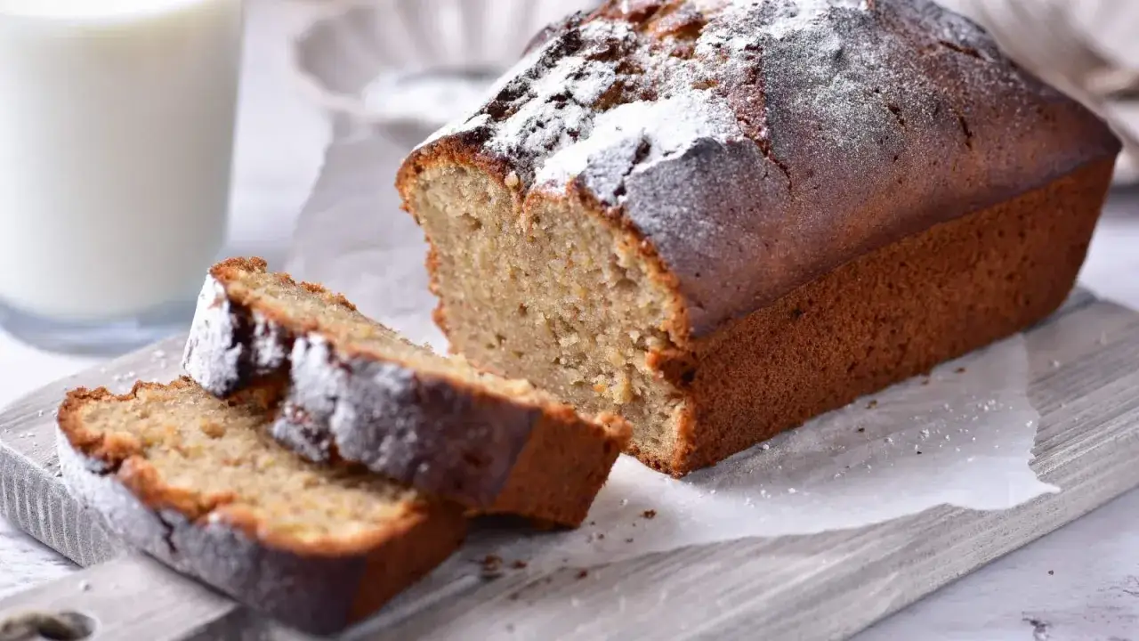 Pyszny chlebek bananowy, oprószony cukrem pudrem, z dwoma kromkami obok. Obok stoi szklanka mleka.