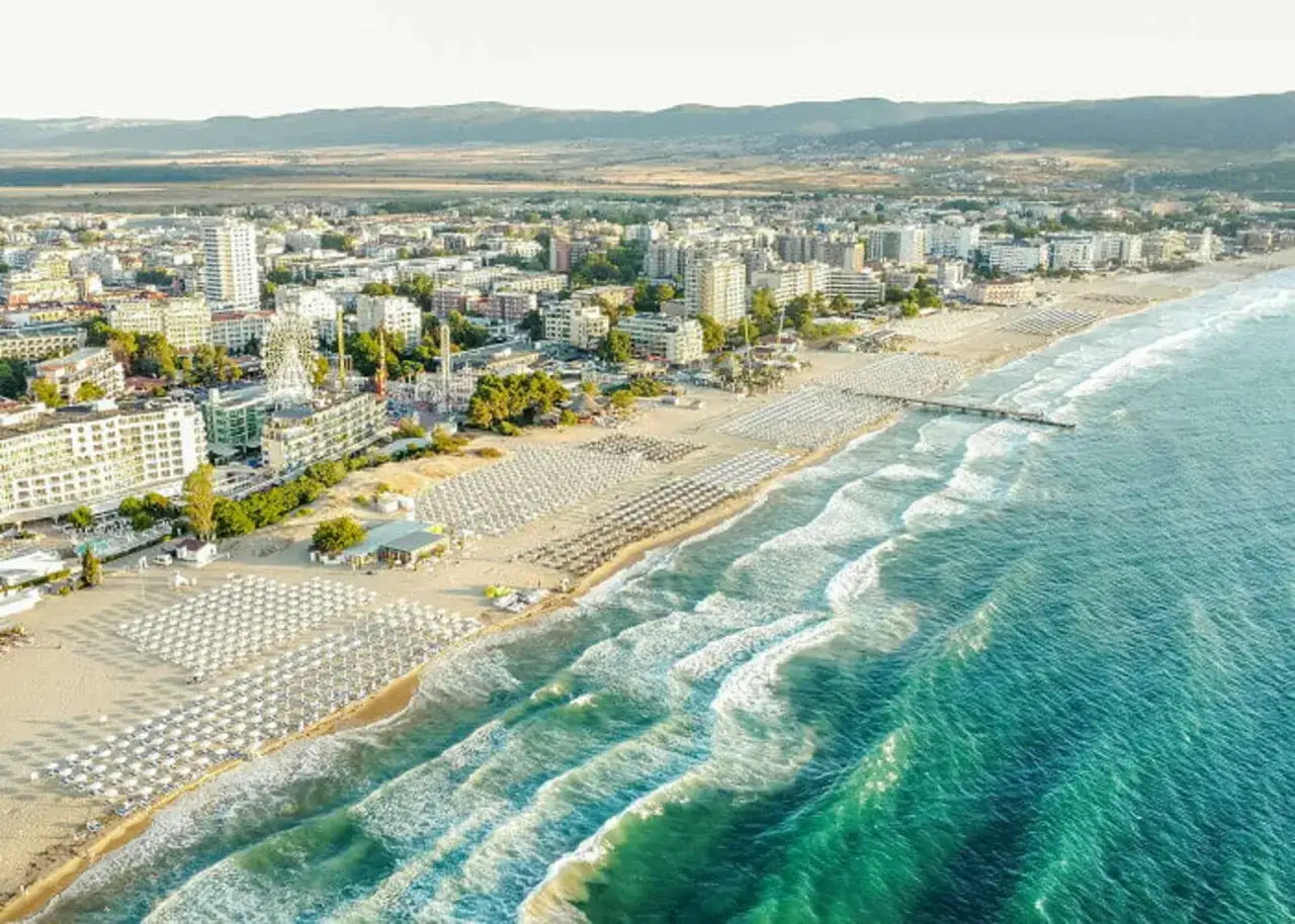 Słoneczna plaża z rzędami leżaków i parasoli, turkusowe morze i miasto w tle. Idealne fajne miejsca na wakacje za granicą.