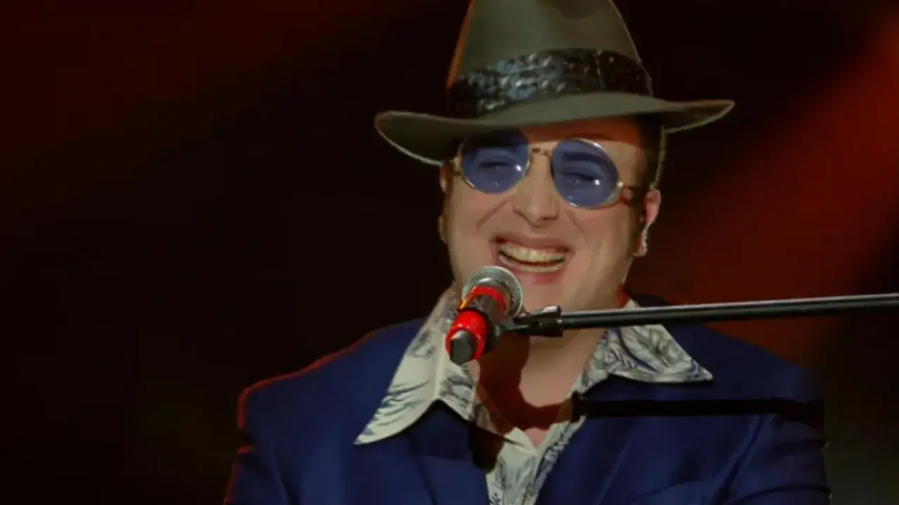 Un artista con cappello e occhiali da sole canta al microfono, un talento tra i cantanti marchigiani.