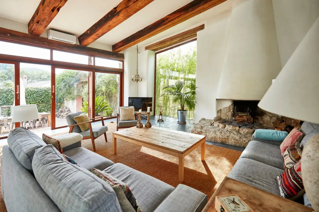 Salón acogedor con chimenea de piedra, vigas de madera y grandes ventanales. Ideal para aprender como encender la chimenea y disfrutar del fuego.