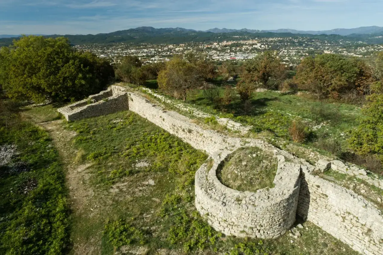 Vestiges de l'oppidum de Jastres, murs de pierre anciens sur une colline verdoyante, avec une ville et des montagnes en arrière-plan.