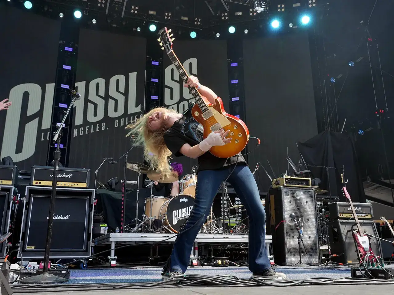 A blonde guitarist with long hair headbangs on stage, playing a sunburst guitar. The band's name, 