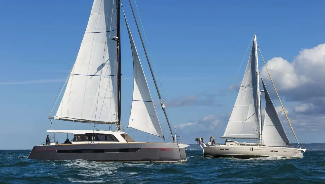 Deux voiliers naviguent sur une mer bleue. L'un est un catamaran gris moderne, l'autre un voilier blanc classique. Une belle journée pour la voile, comme un fruit de mer savoureux sur l'océan.