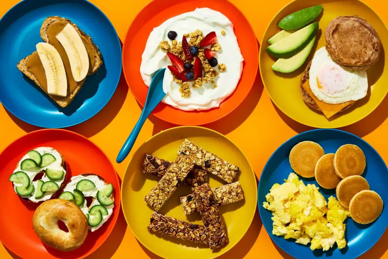 Kolorowe talerze z propozycjami na śniadanie dla rocznego dziecka: kanapka z bananem, jogurt z owocami, jajko sadzone z awokado, bajgle z ogórkiem, batoniki musli, placuszki i jajecznica.