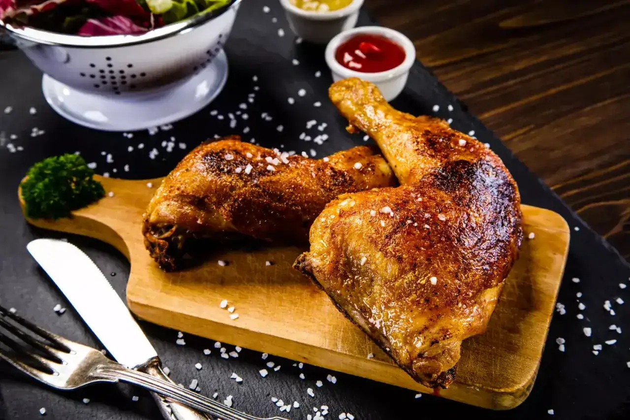 Trois belles cuisses de poulet au four, dorées et croustillantes, servies sur une planche en bois avec une salade et des sauces.