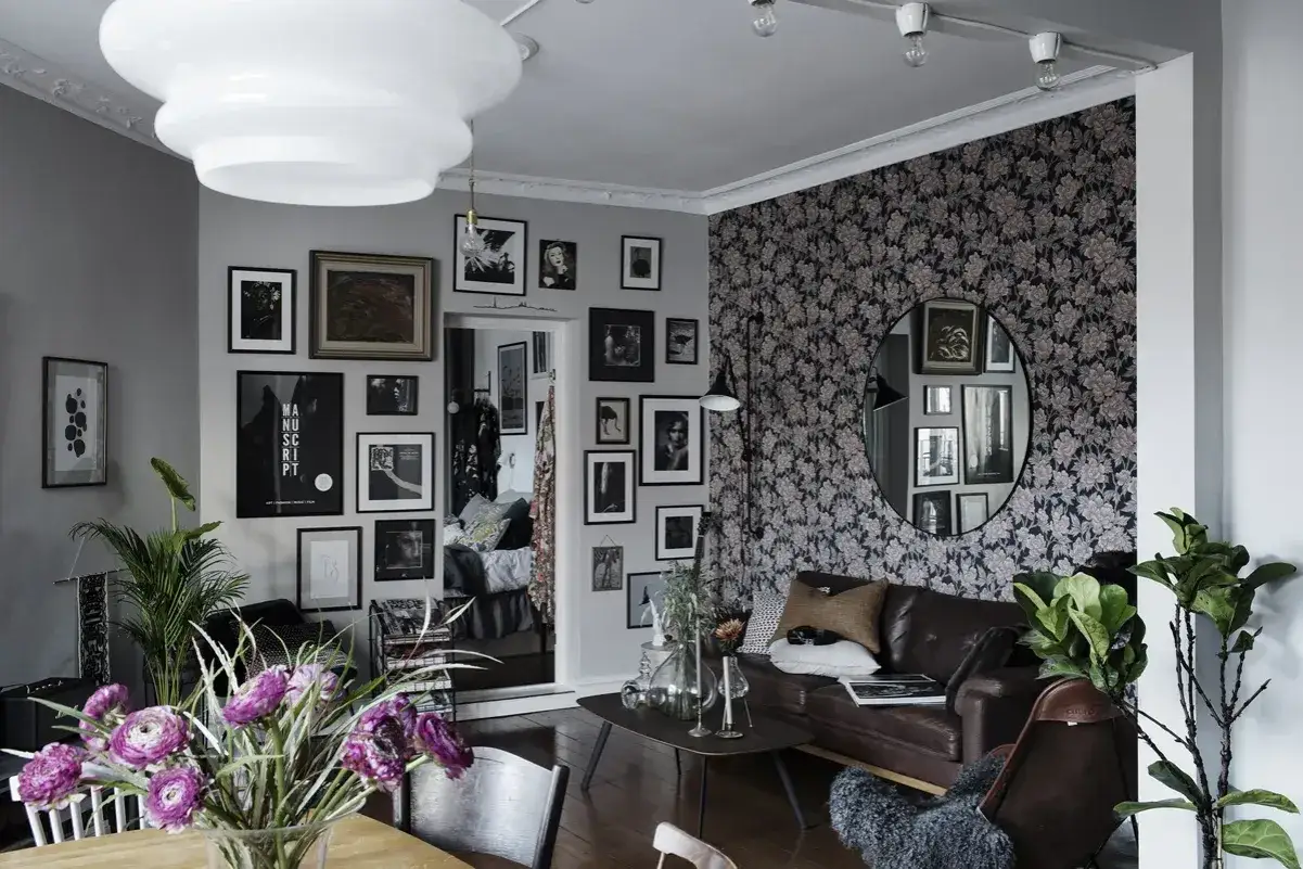 Ett vardagsrum i bohemisk stil med en v&auml;gg t&auml;ckt av blommig tapet, en l&auml;dersoffa, m&aring;nga tavlor och en stor rund spegel.