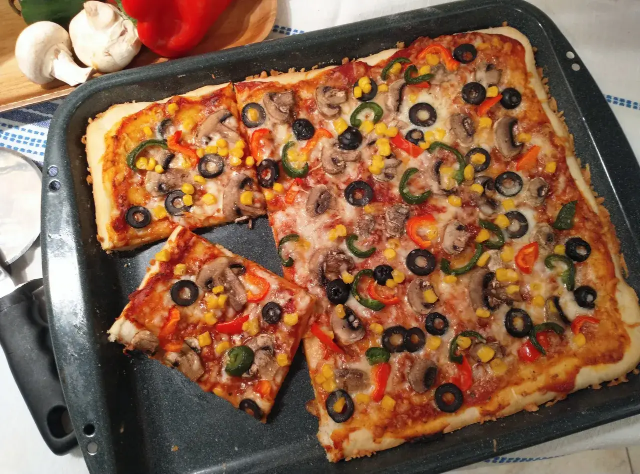 Pyszna pizza bez mięsa z pieczarkami, kukurydzą, papryką i oliwkami na prostokątnej blasze.