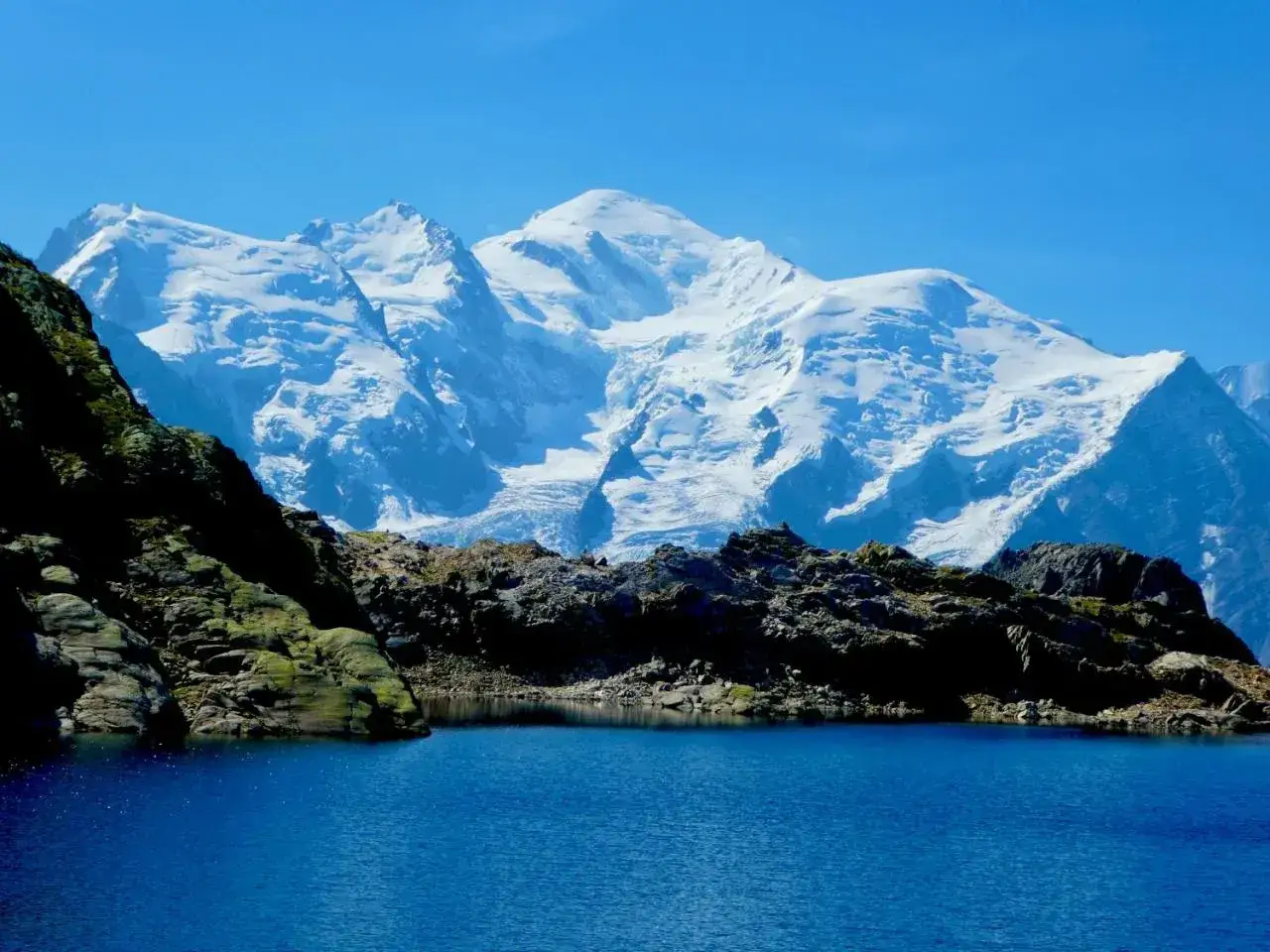 Lac d'altitude aux eaux bleues, refl&eacute;tant les sommets enneig&eacute;s du tour des Aiguilles Rouges sous un ciel clair.