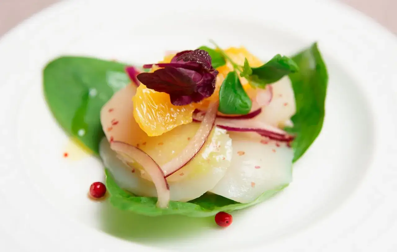 Carpaccio de Saint-Jacques aux agrumes, oignons rouges et baies roses sur une feuille verte.