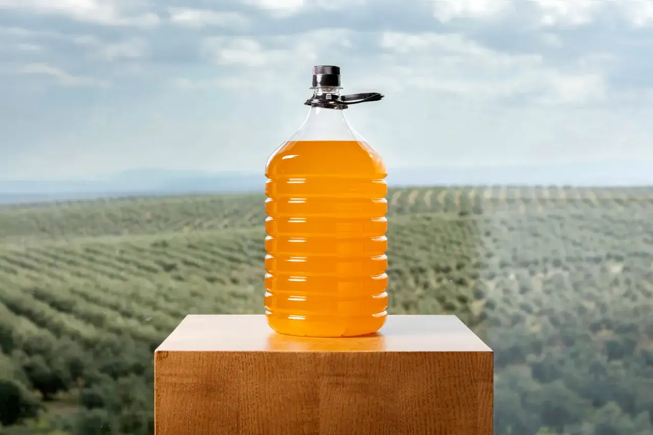 Botella grande de aceite de oliva lampante, de color naranja intenso, sobre un pedestal de madera con un campo de olivos al fondo.