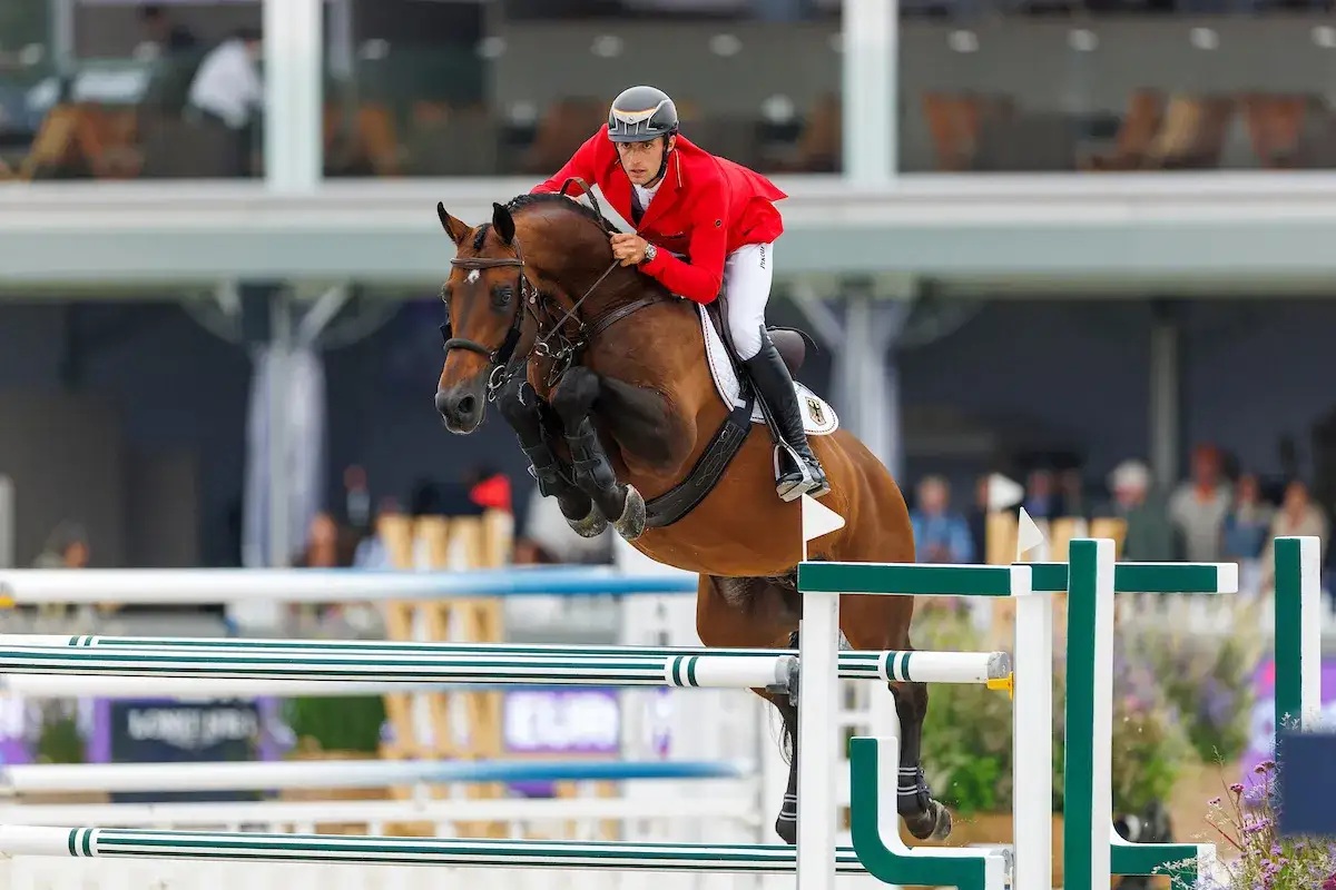 Reiter in rotem Sakko springt mit braunem Pferd über Hindernis bei einem spannenden **Turnier Reiten**.