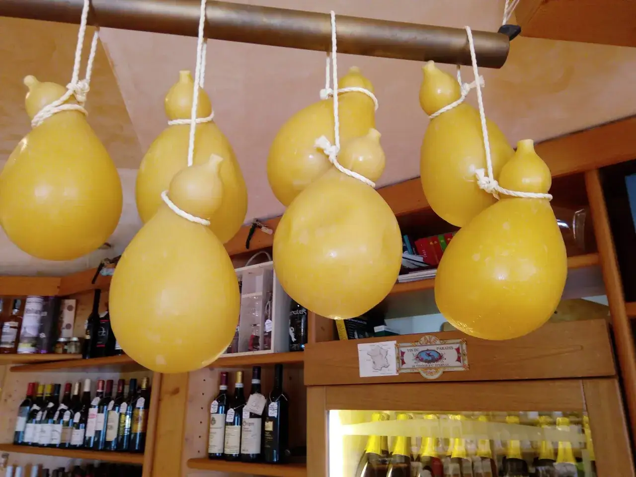 Formaggi podolici appesi a stagionare, con bottiglie di vino sugli scaffali sottostanti.