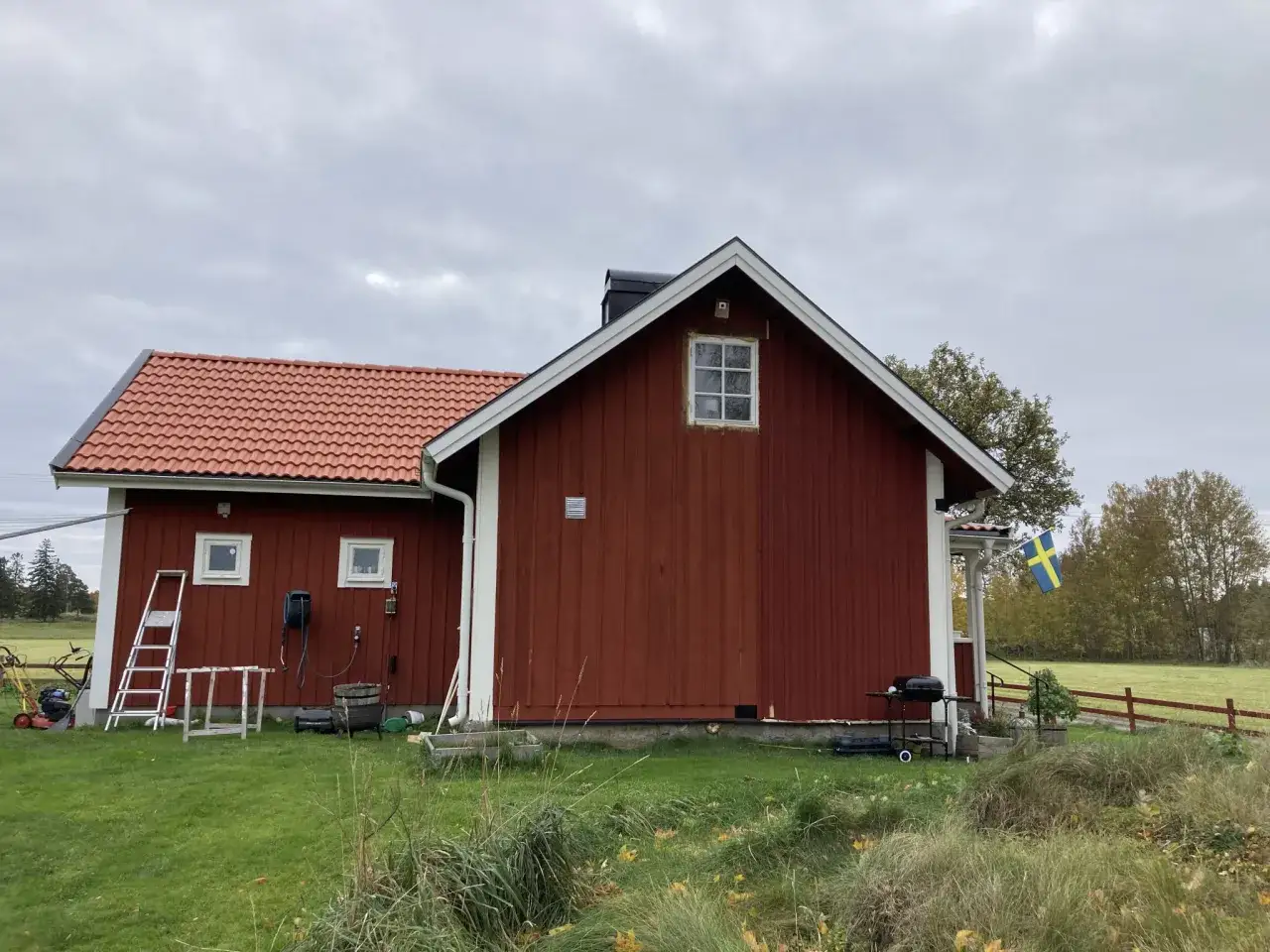 Rött torp med ny tillbyggnad, svensk flagga vajar. Gräsklippare och grill står redo.