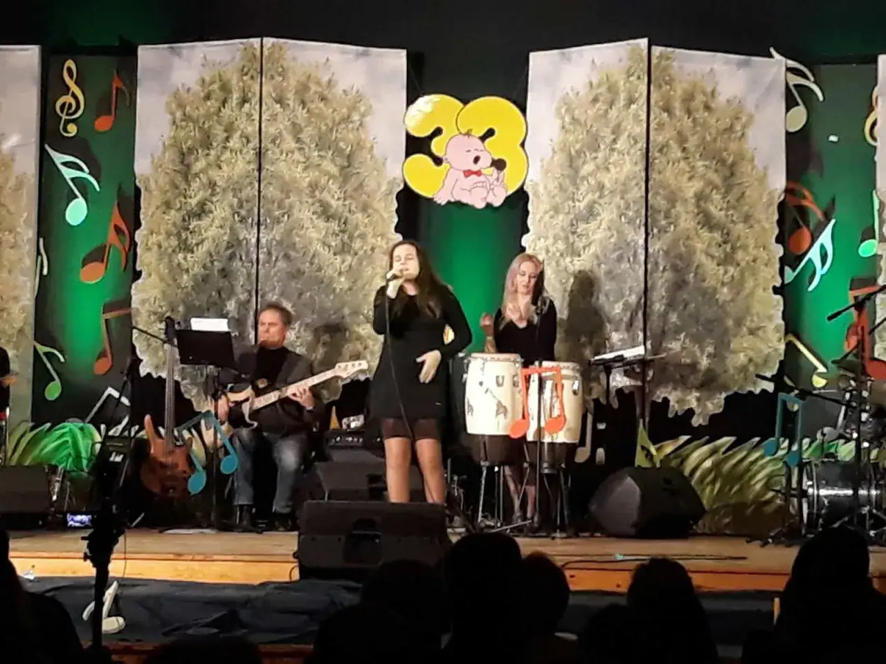 Zespół muzyczny na scenie. Wokalistka, gitarzysta i perkusistka. Scenografia z drzewami i nutami. Ciekawe, jakie wykształcenie muzyczne mają najlepsi zawodnicy w historii teleturnieju.