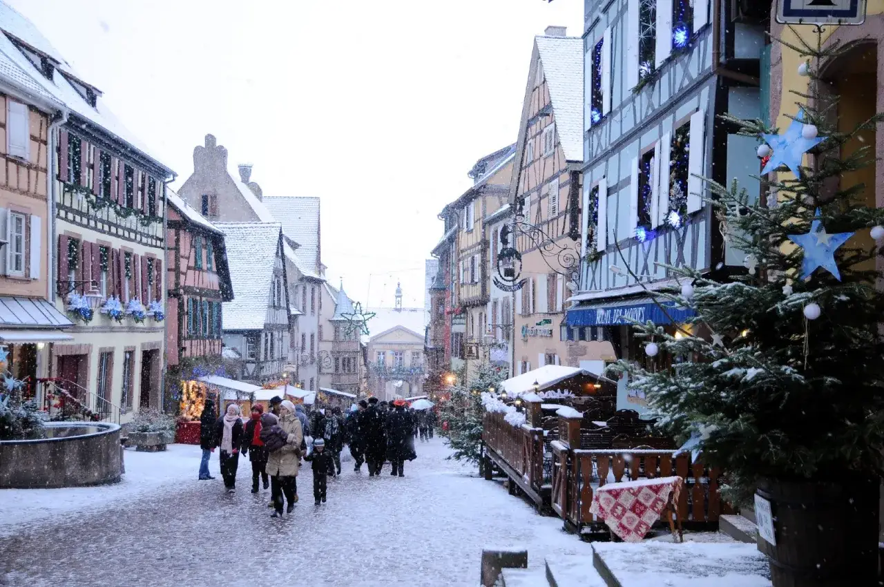 Un pueblo franc&eacute;s en invierno, con casas de entramado de madera cubiertas de nieve y gente paseando entre puestos navide&ntilde;os.