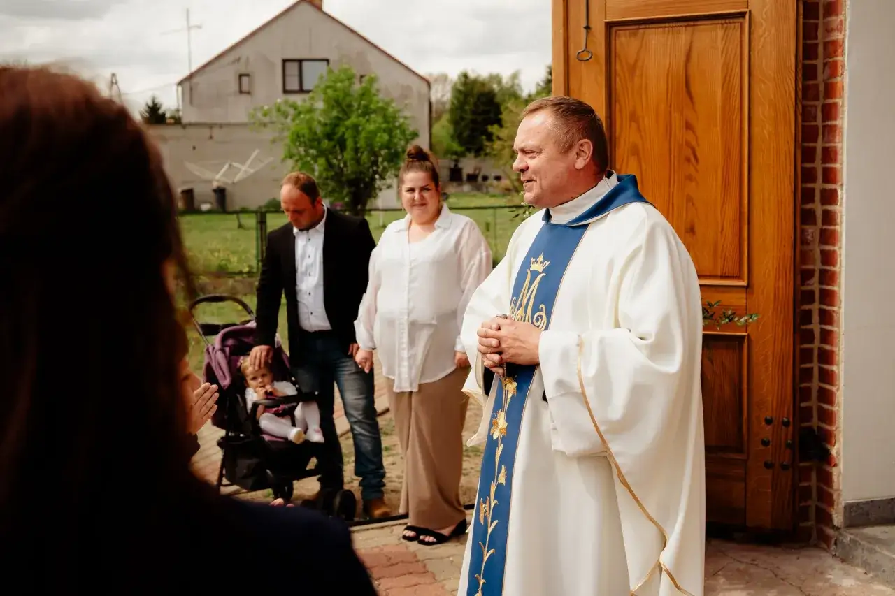 Kiedy są komunie, ksiądz w białej albie z niebieską stułą błogosławi rodzinę z dzieckiem w wózku przed kościołem.
