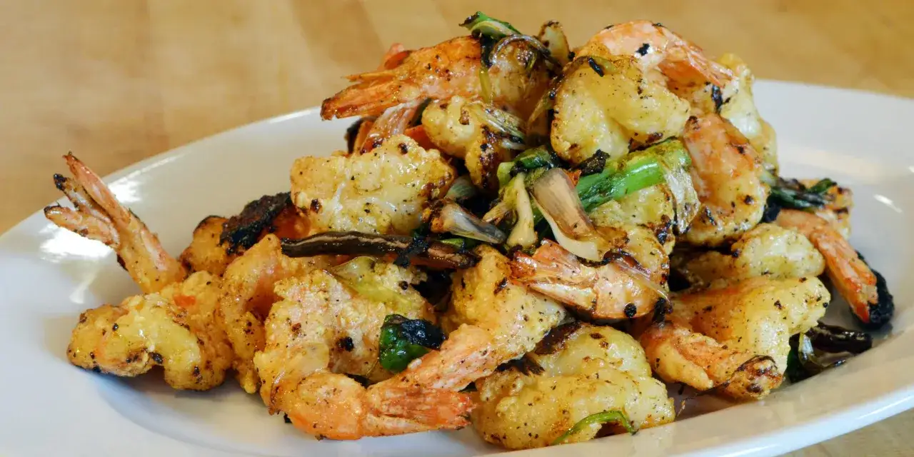 Gamberi sale e pepe croccanti, dorati e conditi con erbe aromatiche, serviti su un piatto bianco.