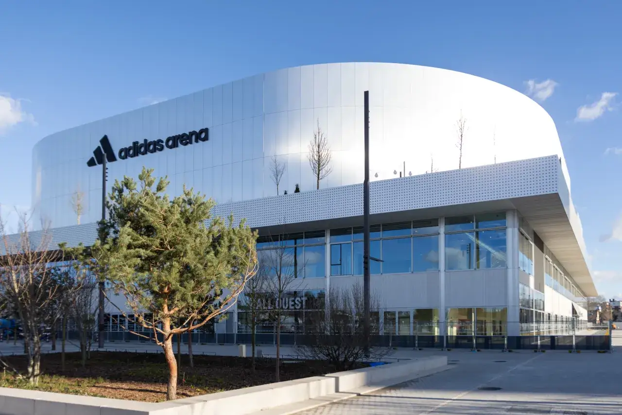 La moderne adidas arena Paris, avec son logo emblématique, se dresse sous un ciel bleu. Des arbres verdoyants bordent l'entrée.