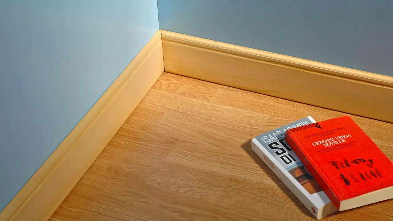 Angolo di una stanza con parquet e pareti azzurre. La posa in opera battiscopa in legno è curata. Due libri giacciono sul pavimento.