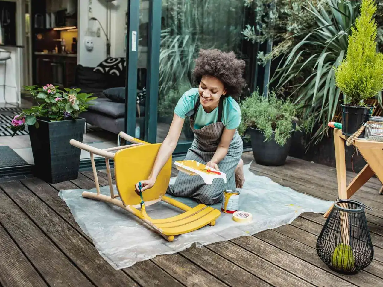 Une femme souriante est à genoux sur une terrasse en bois, occupée à peindre un meuble en bois jaune vif.