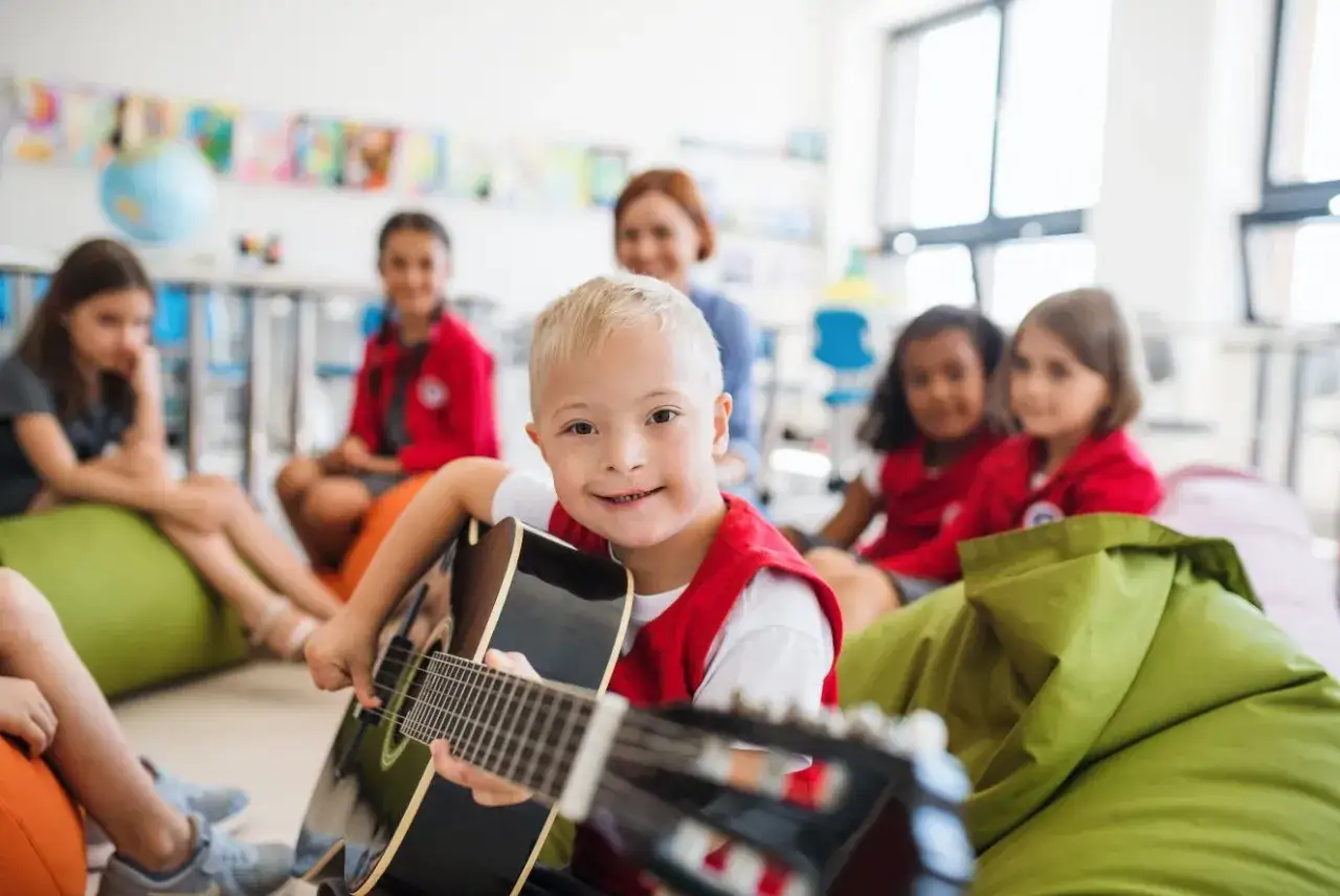 Ein Junge mit Down-Syndrom spielt Gitarre in einer Klasse. Die Lehrerin und andere Kinder schauen zu. Die Musiktherapie hilft, die Entwicklung der Kinder zu fördern.