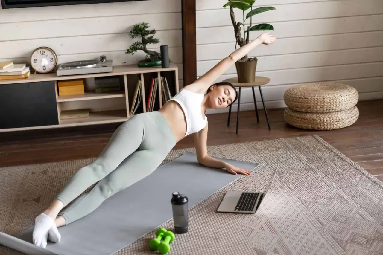 Mujer en postura de plancha lateral, disfrutando de los beneficios del yoga en casa. A su lado, un port&aacute;til y pesas verdes.