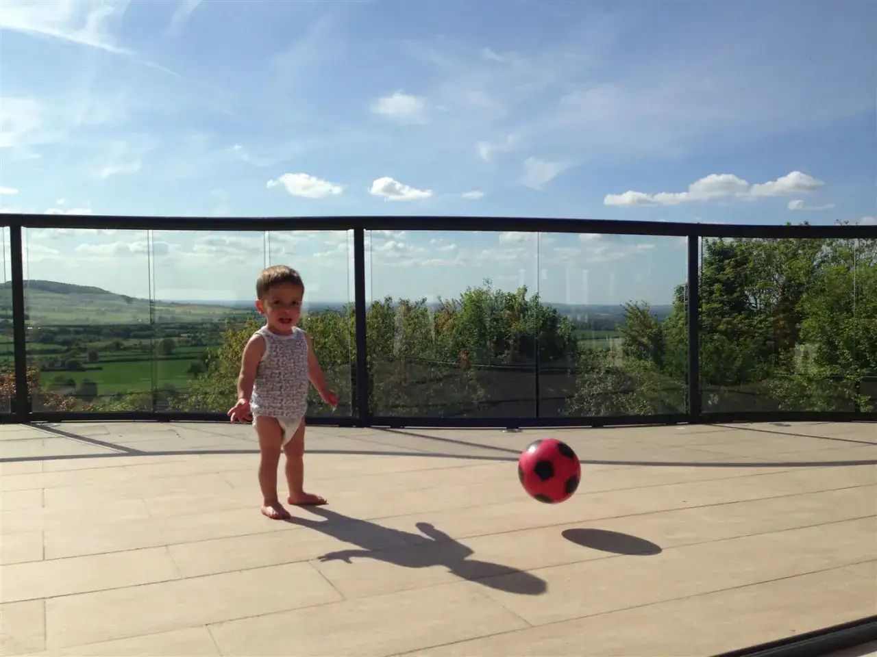 Kleines Kind spielt auf einem kindersicheren Balkon mit einem roten Fußball. Die Aussicht ist ins Grüne.
