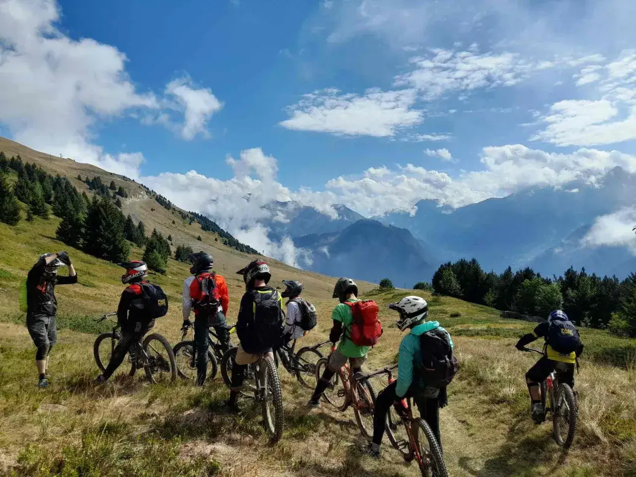 Groupe d'ados en colonie VTT descente, prêts à dévaler les pentes montagneuses sous un ciel bleu et nuageux.