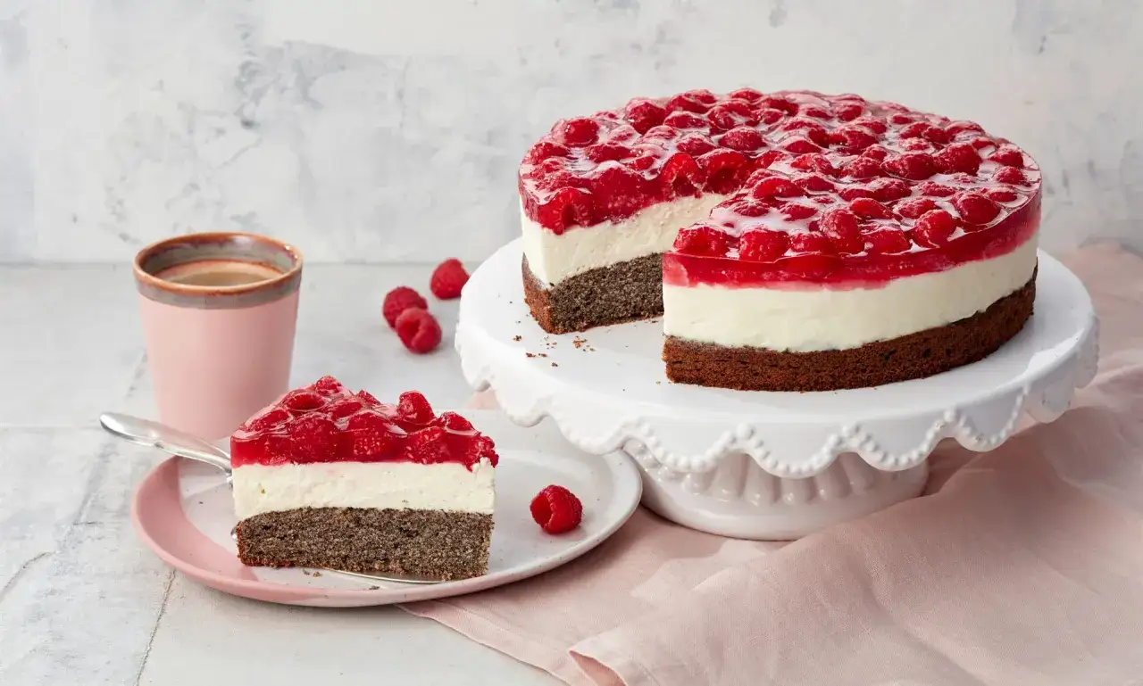 Himbeer Mohn Torte mit einer Schicht aus wei&szlig;er Creme und einem Mohnboden, bedeckt mit frischen Himbeeren und Gelee. Ein St&uuml;ck ist auf einem Teller angerichtet.
