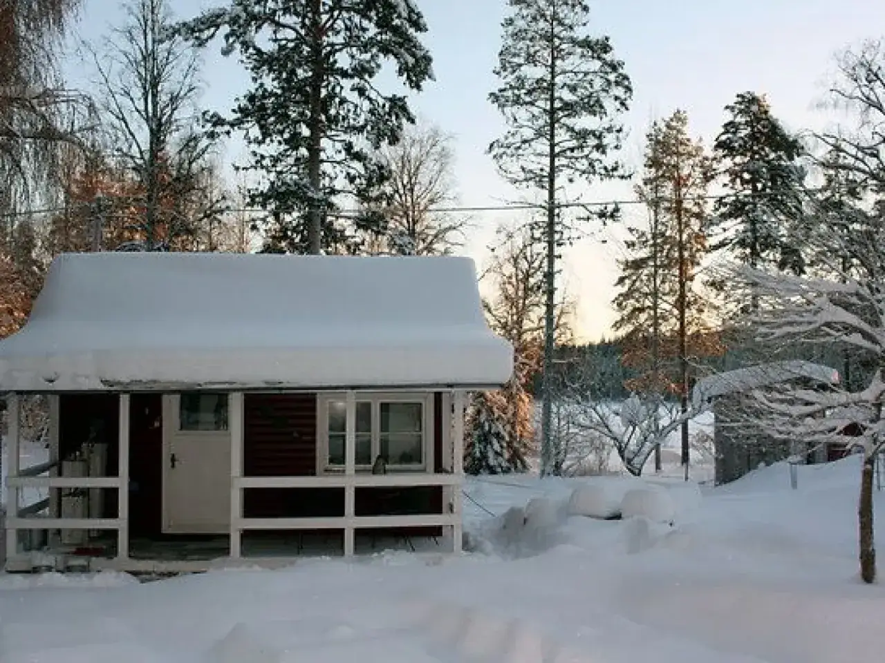 Snötäckt stuga med en veranda. Inne i stugan syns ett fotogenkök, redo för en mysig vinterkväll.