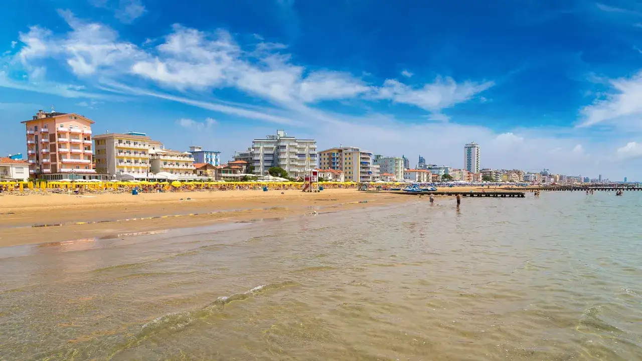 Spiaggia affollata con ombrelloni gialli e hotel colorati sul lungomare del mare veneto. Acqua limpida e cielo azzurro con nuvole bianche.