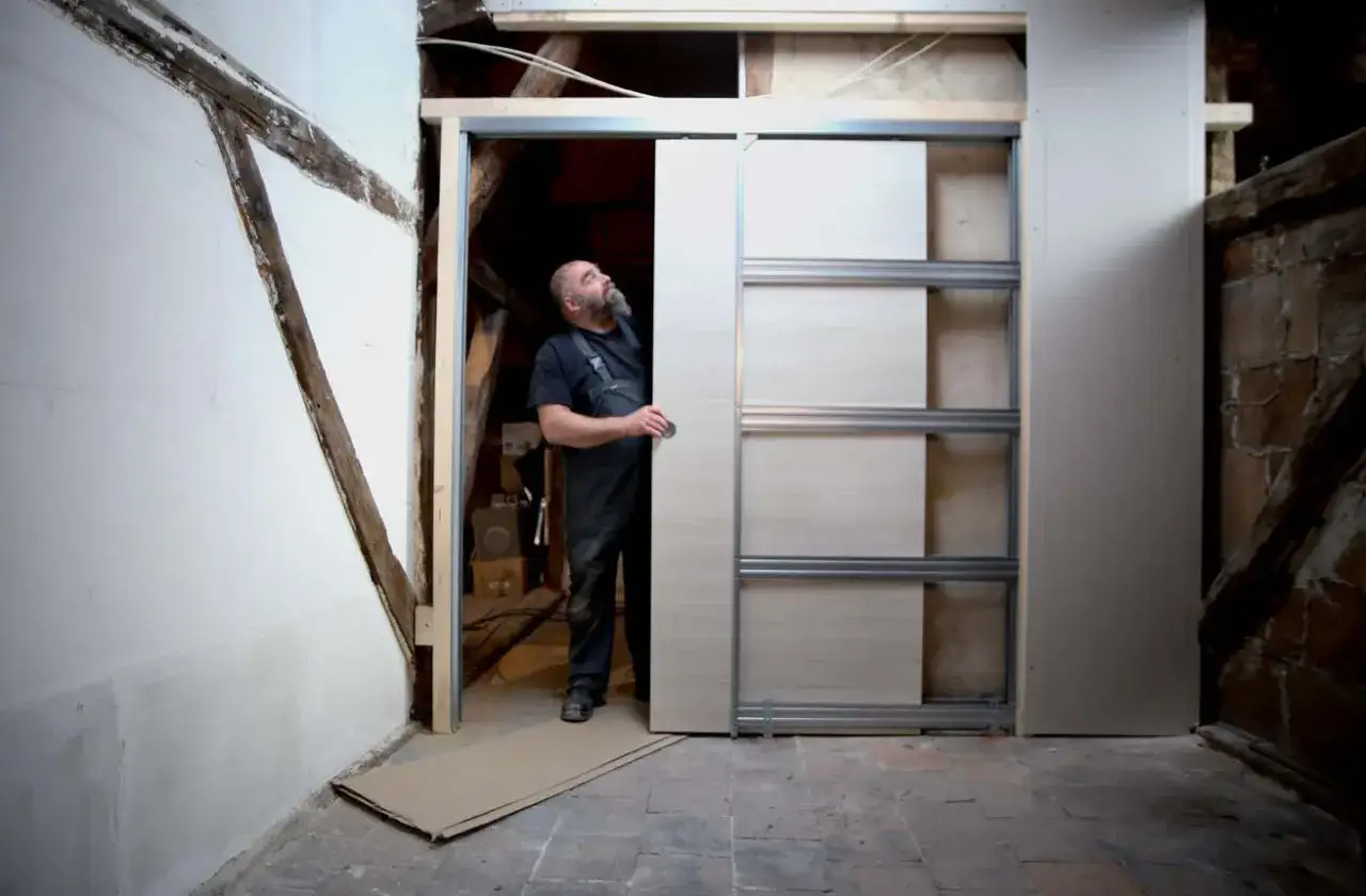 Un homme installe une porte coulissante dans une pièce en rénovation. La structure métallique est visible.
